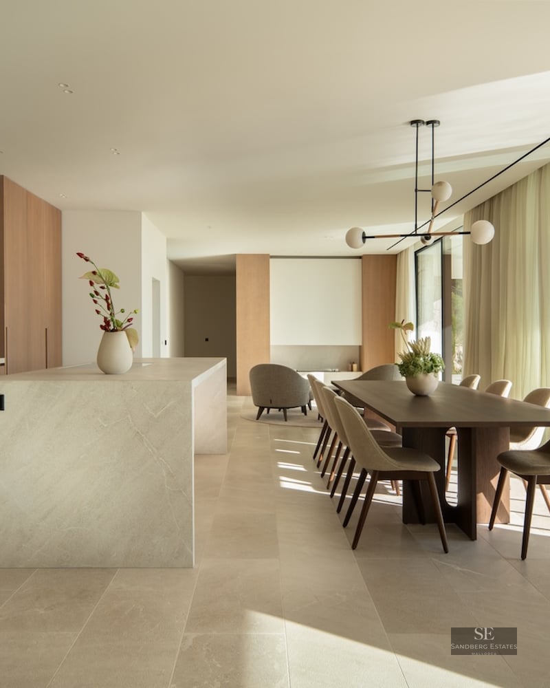 Modern dining room with a wooden table, stone kitchen island, and large floor-to-ceiling windows overlooking a forest.