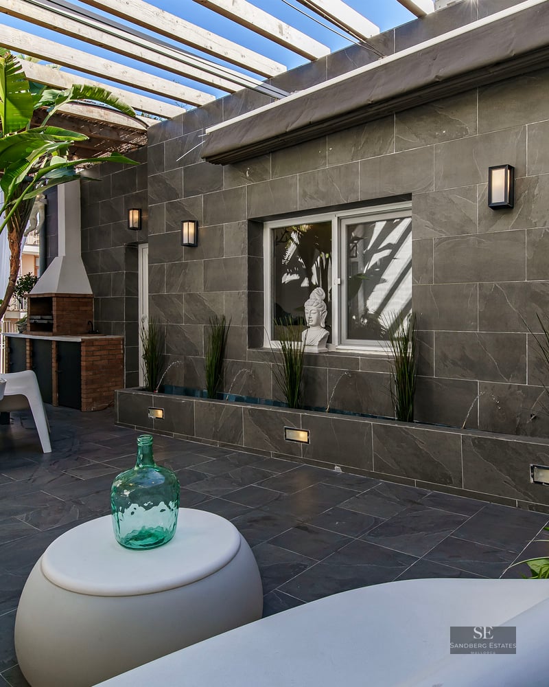Luxury patio with grey stone walls, white modern furniture, a wooden pergola, and tropical plants.