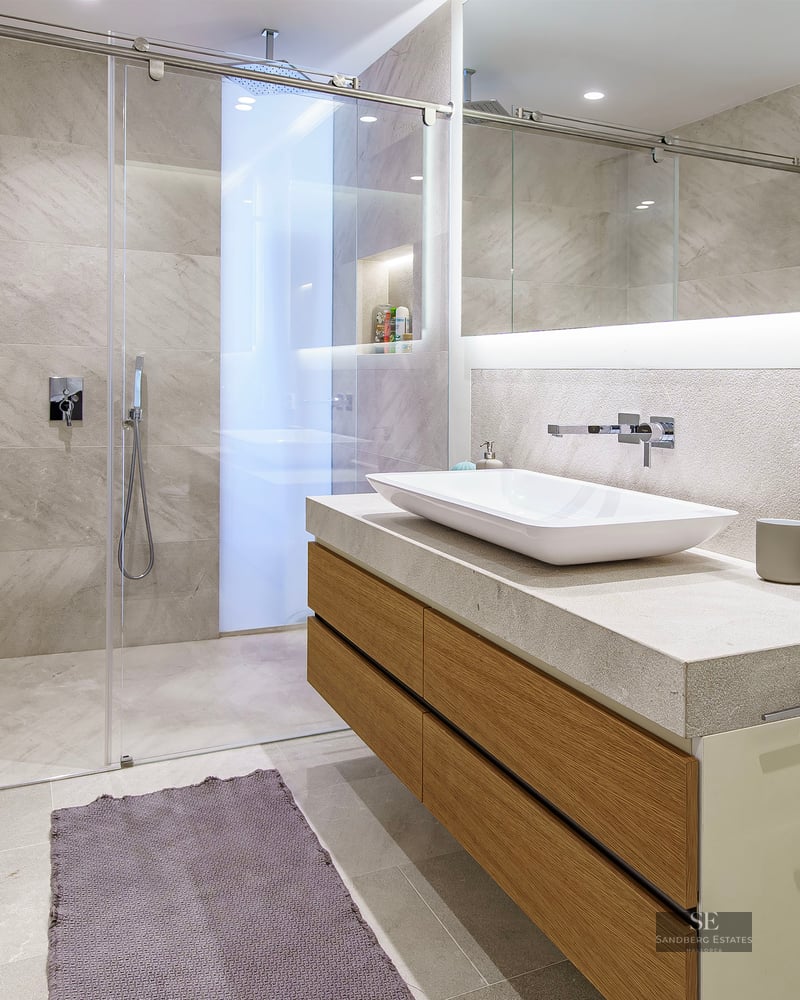 Salle de bain moderne avec douche en verre, meuble en bois avec vasque blanche et grand miroir rétroéclairé.
