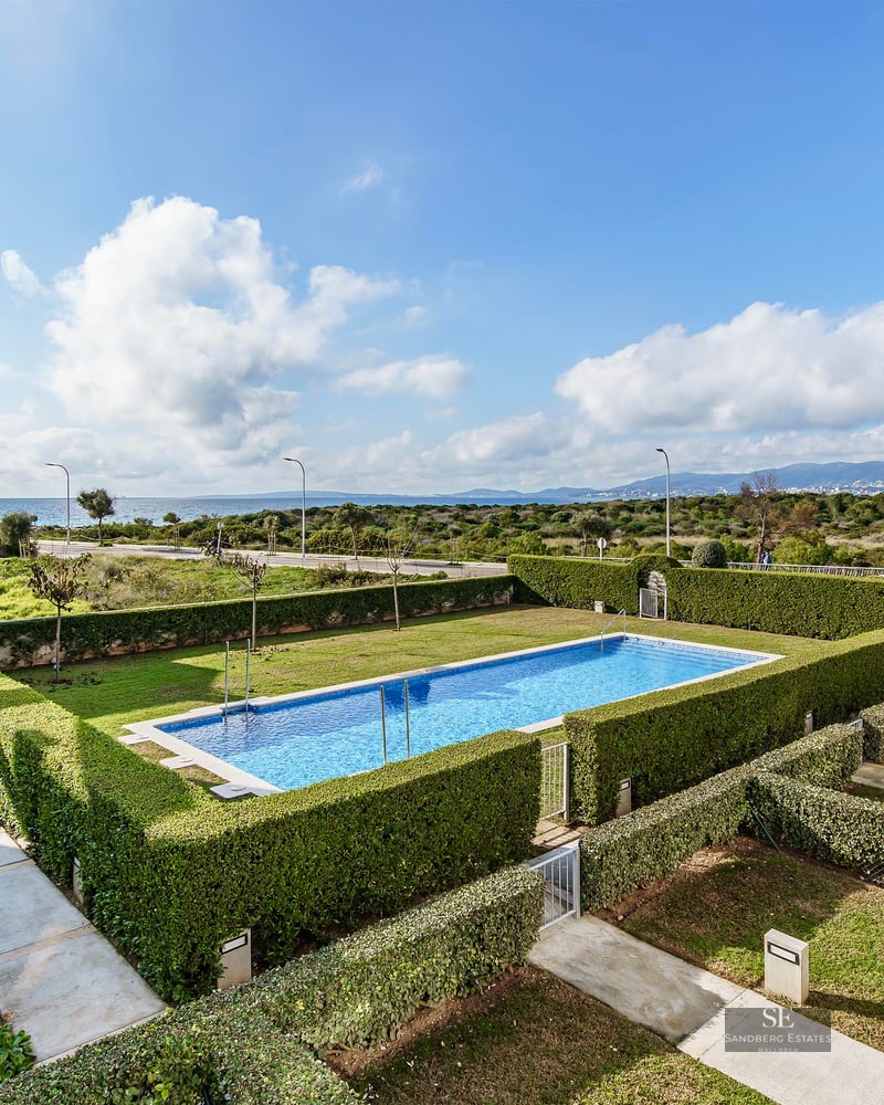 Rectangular blue swimming pool surrounded by manicured green hedges with a view of the sea in the distance.