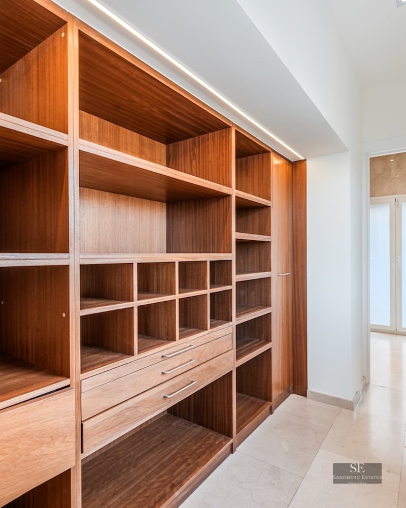 Modern walk-in closet with warm wood shelving, marble floors, and integrated LED lighting next to a bright window.