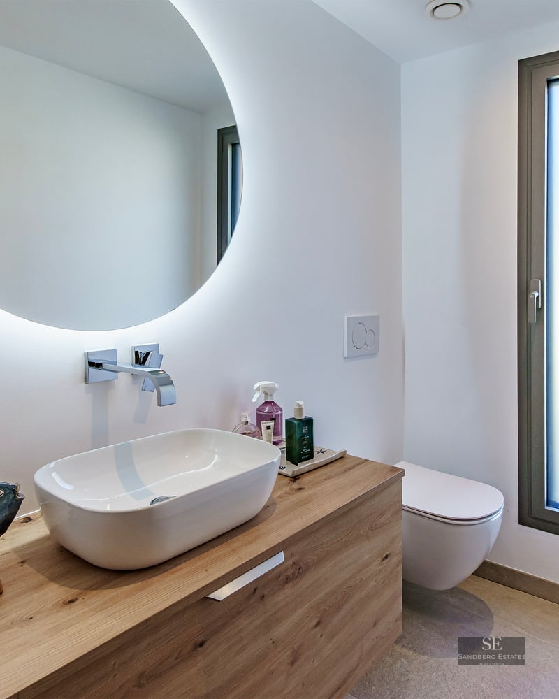 Modern bathroom featuring a white vessel sink on a wooden vanity, a large round backlit mirror, and a frosted glass window.