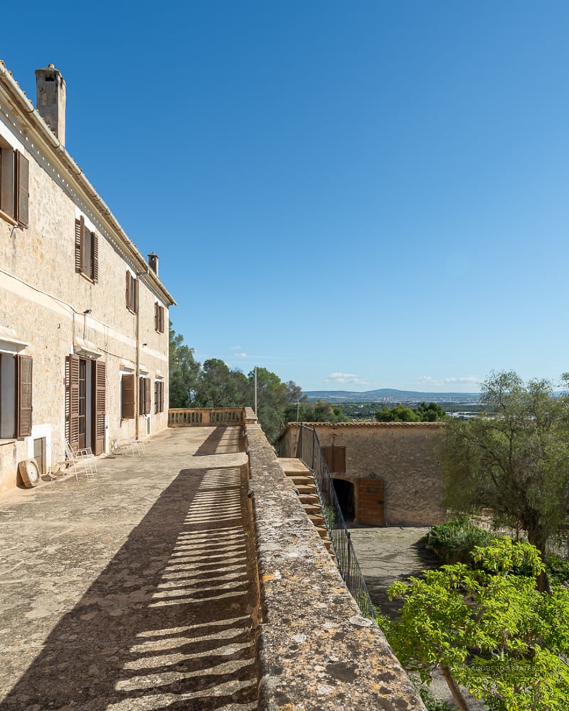 A long stone terrace on a rustic building with wooden shutters, overlooking a green landscape under a clear blue sky.