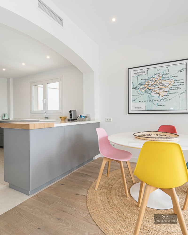 Salle à manger lumineuse avec table ronde blanche, chaises colorées et tapis en jute à côté d'une cuisine grise.