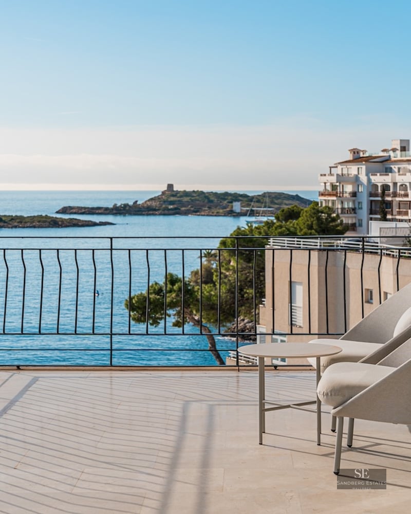 Modern balcony with grey armchairs overlooking a sparkling blue sea with islands and coastal buildings.