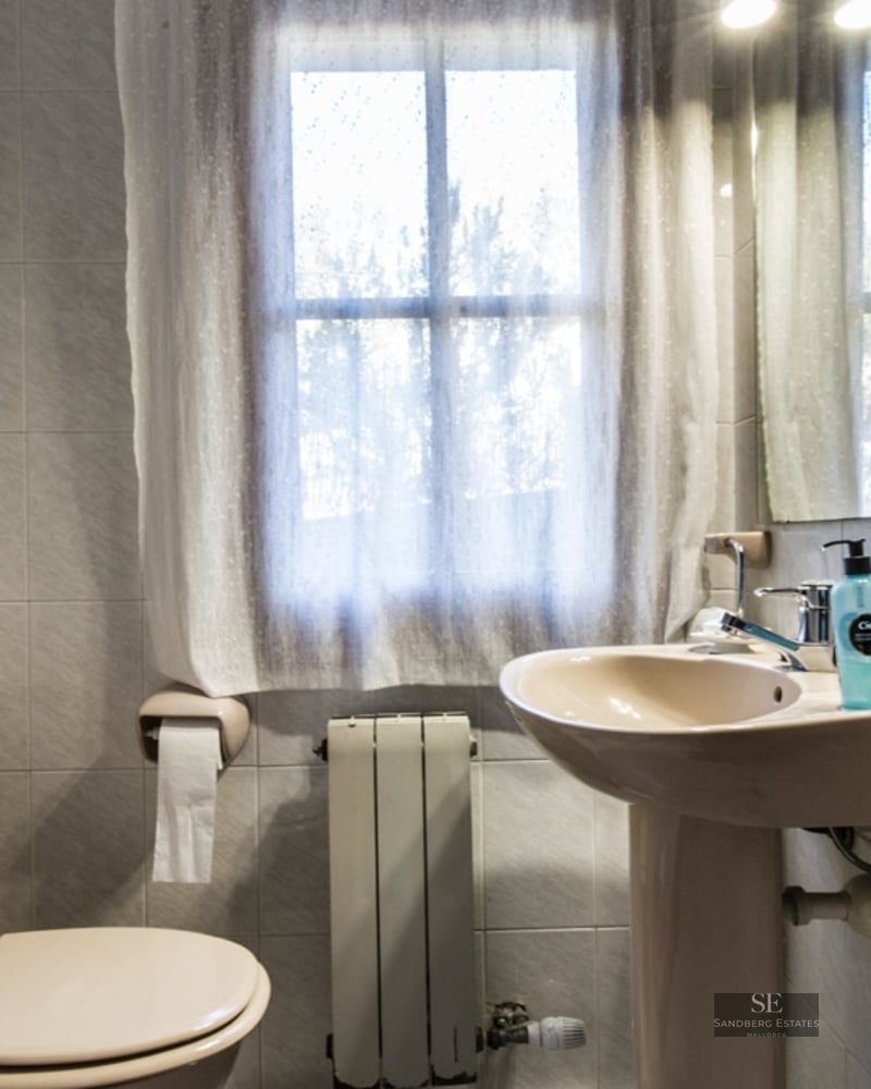 Small bathroom featuring a beige toilet and pedestal sink, a window with a sheer curtain, and light-tiled walls.