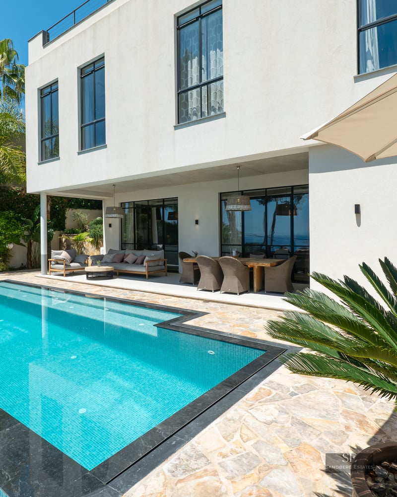 Bright outdoor scene featuring a turquoise infinity pool, covered terrace, and modern white villa with palm trees.