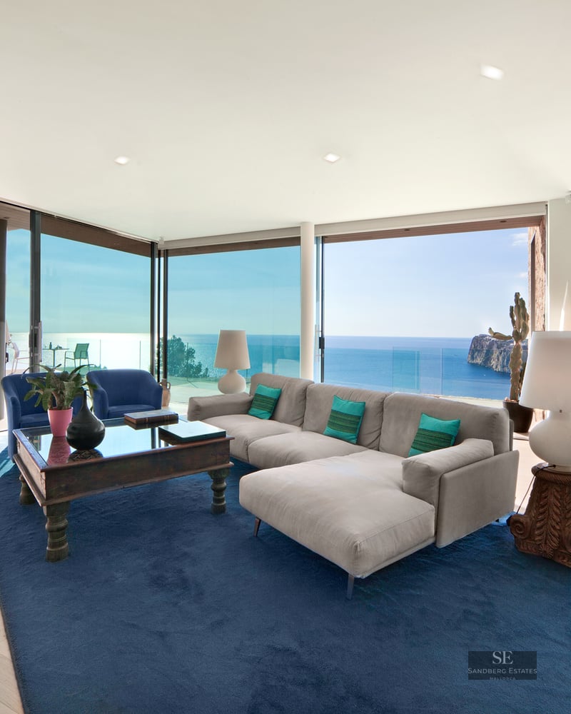 Modern living room featuring a gray sectional sofa, large blue rug, and floor-to-ceiling windows overlooking the ocean.