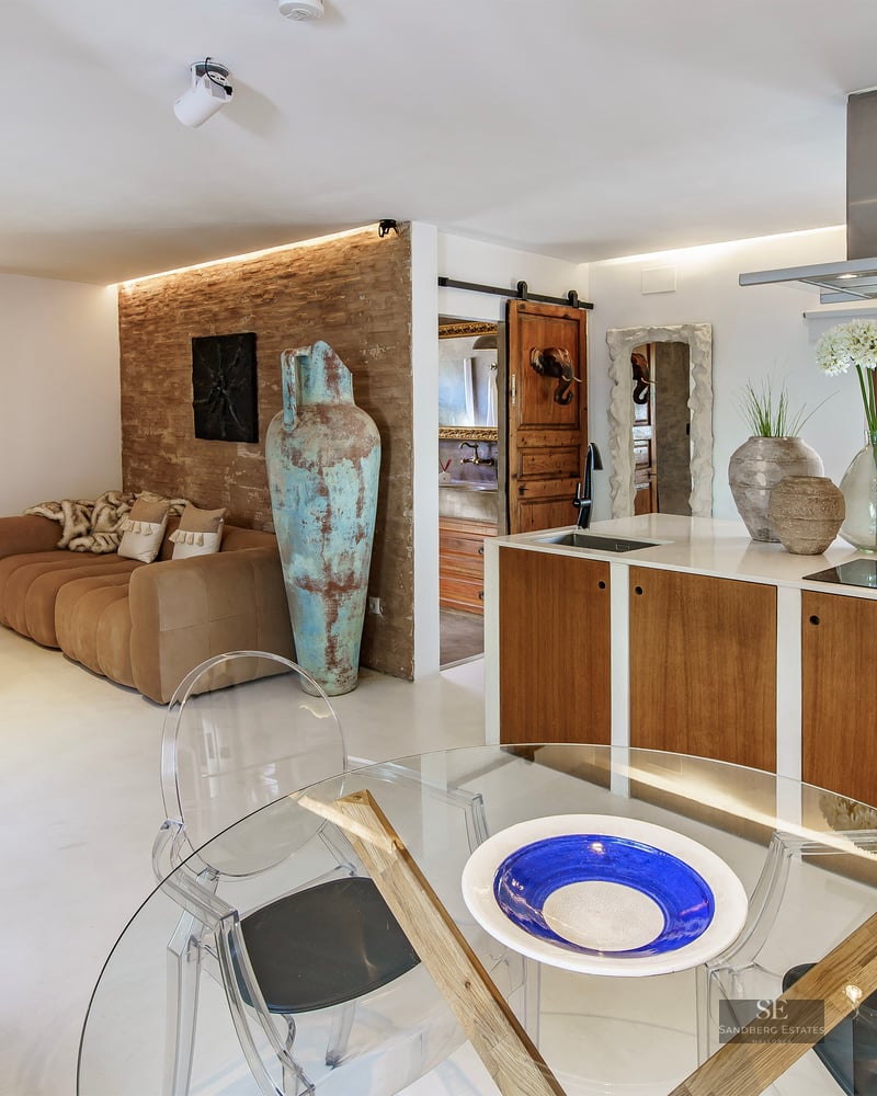 Open-plan living room and kitchen featuring a brown sofa against a brick wall, a large turquoise vase, and glass table.