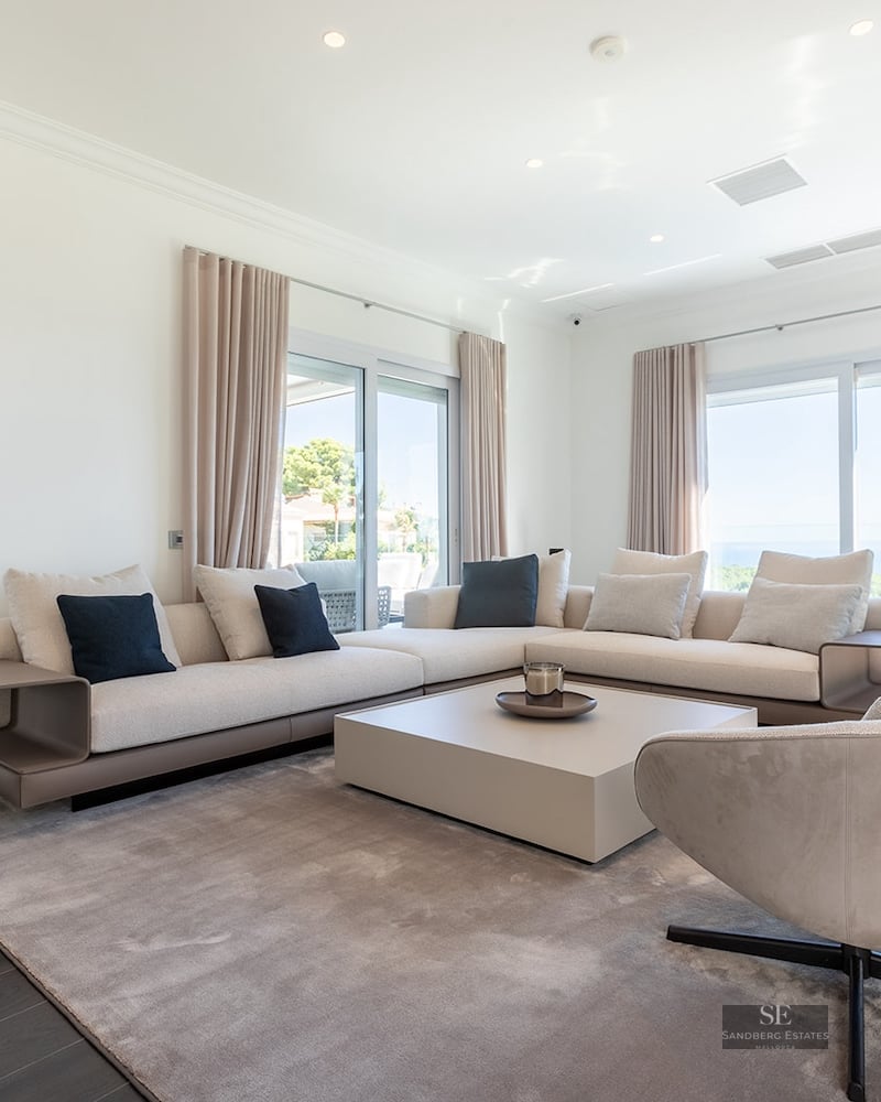 Spacious white living room with beige sectional sofa, designer armchair, and large windows overlooking the ocean.