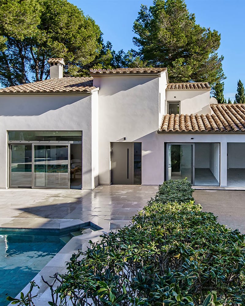 Modern white villa with a turquoise swimming pool, stone terrace, and lush pine trees under a clear blue sky.
