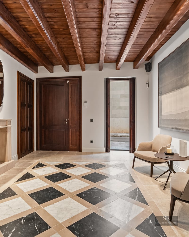 Luxury entry with marble checkerboard floor, exposed wood ceiling beams, stone fireplace, and designer armchairs.