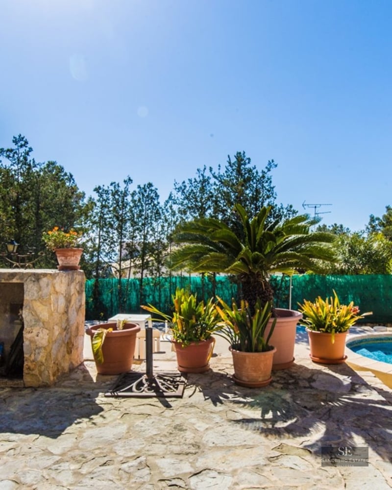 Sun-drenched stone terrace featuring a blue swimming pool, built-in stone barbecue, and lush potted plants under a clear sky.