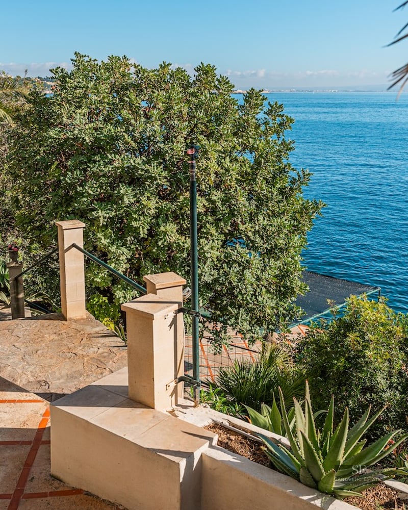 A sunny stone terrace with terracotta tiles leads to a lush garden with palm trees overlooking the blue ocean.