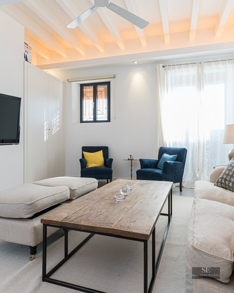 Bright living room with white beamed ceiling, beige sofa with colorful cushions, and wall-mounted flat-screen TV.