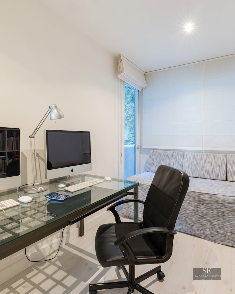 A bright home office featuring a glass desk with two computers, a black ergonomic chair, and floor-to-ceiling bookshelves.