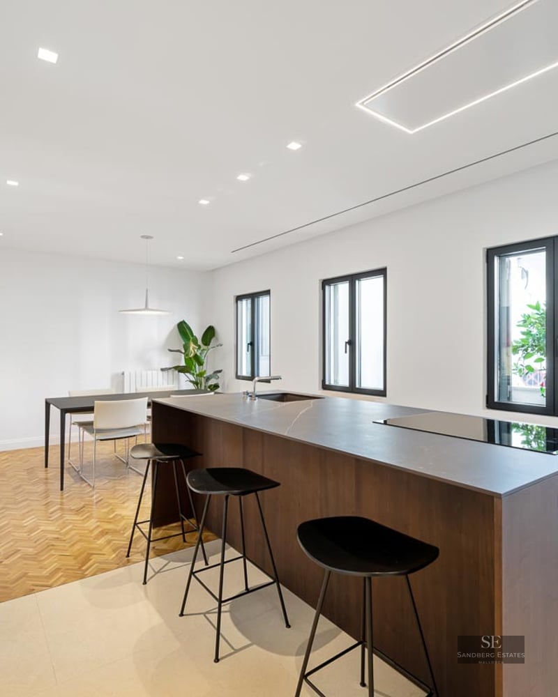 Minimalist kitchen featuring a dark wood island, black bar stools, herringbone wood flooring, and abstract wall art.