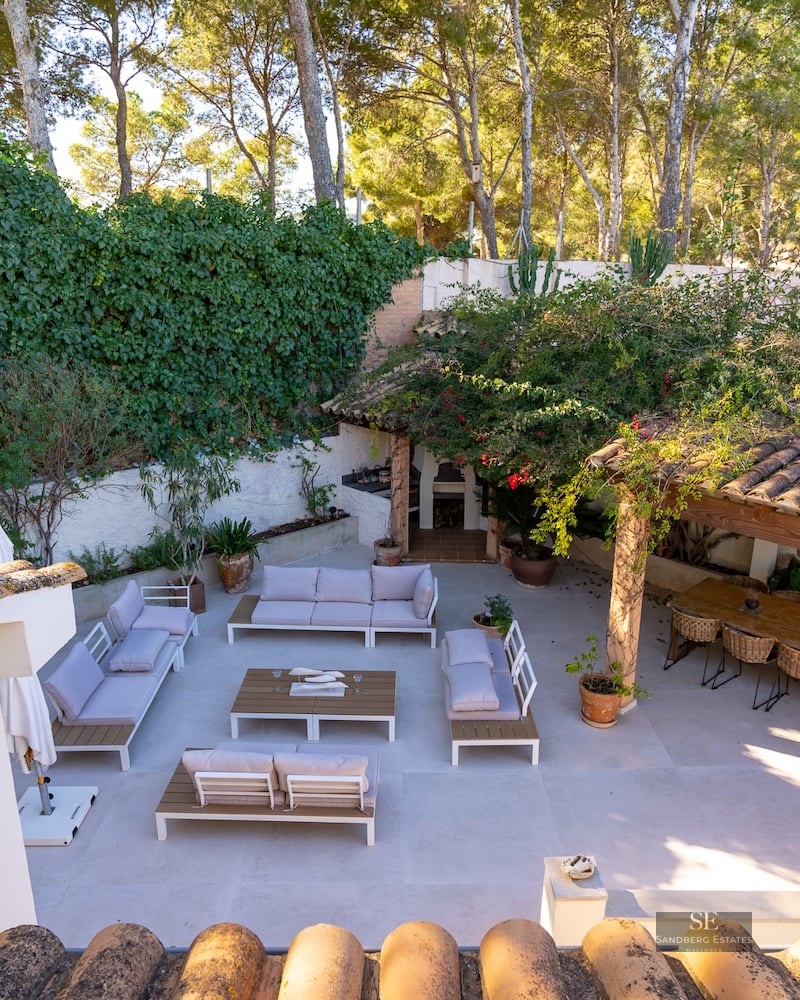 Elevated view of a spacious paved terrace with white lounge sofas, a large wooden dining table, and lush green walls.