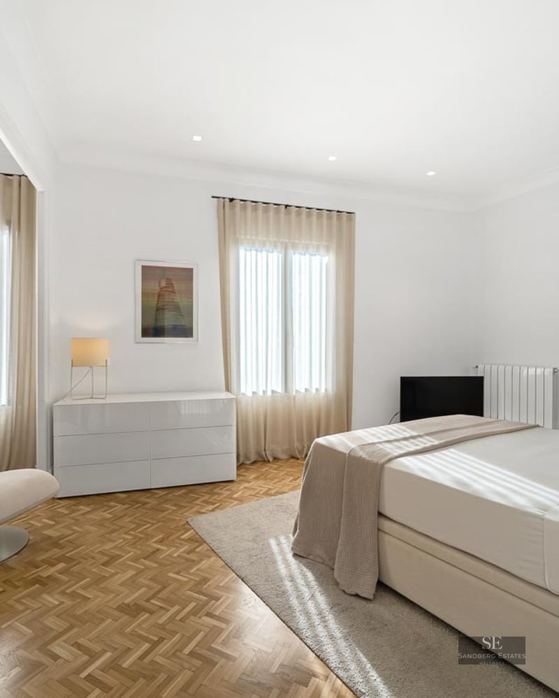 Bright bedroom with white walls, herringbone parquet, a double bed, designer chair, and sheer curtains.