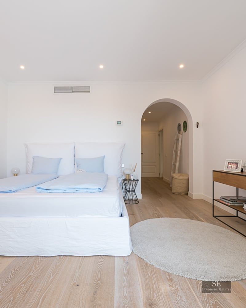 Chambre lumineuse avec murs blancs, parquet clair, grand lit avec draps bleus et passage voûté vers le couloir.