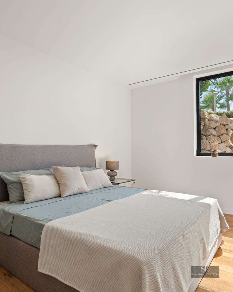 Bright modern bedroom with a gray upholstered bed, glass nightstands, wooden floors, and a large window.