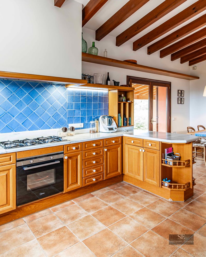 Rustic kitchen featuring wooden cabinets, blue tiled backsplash, terracotta floors, and exposed ceiling beams.