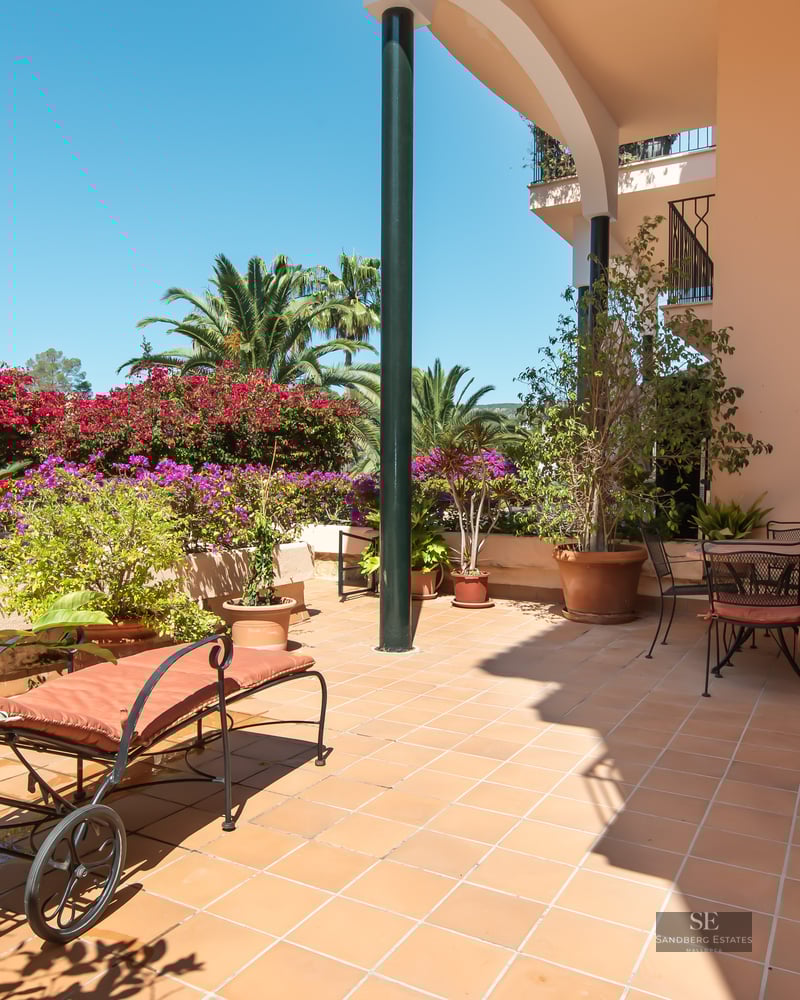 Terracotta terrace with a sun lounger and dining area surrounded by bougainvillea and palm trees under a blue sky.