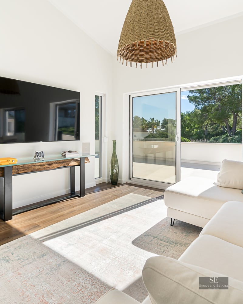 Salon moderne avec canapé d'angle blanc, TV murale, console rustique et grandes baies vitrées coulissantes.