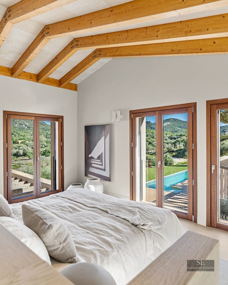 Modern bedroom with vaulted wood beam ceiling, bed with beige linens, and glass doors overlooking a pool and green hills.
