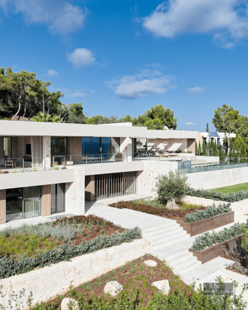 Modern two-story luxury villa with stone facades, glass balconies, and tiered Mediterranean gardens overlooking the sea.