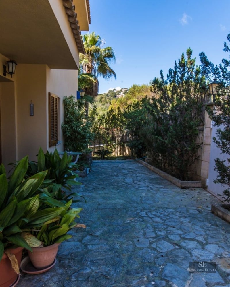 Stone-paved garden pathway alongside a beige Mediterranean house with lush green plants and terracotta pots.