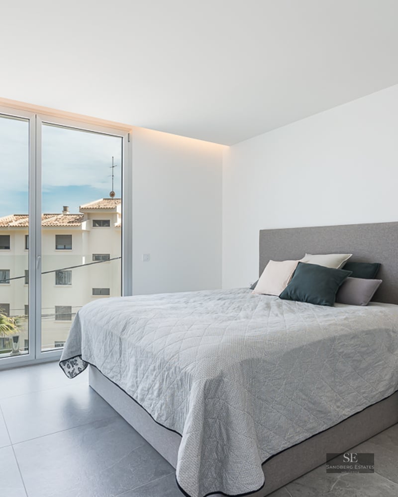 Bright modern bedroom with a large window overlooking a coastal town and sea, featuring a grey bed and tiled floor.