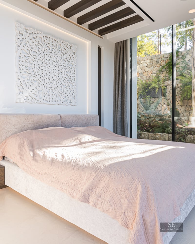 Bright master bedroom with a large bed, intricate wall art, and floor-to-ceiling windows looking out to a stone garden wall.