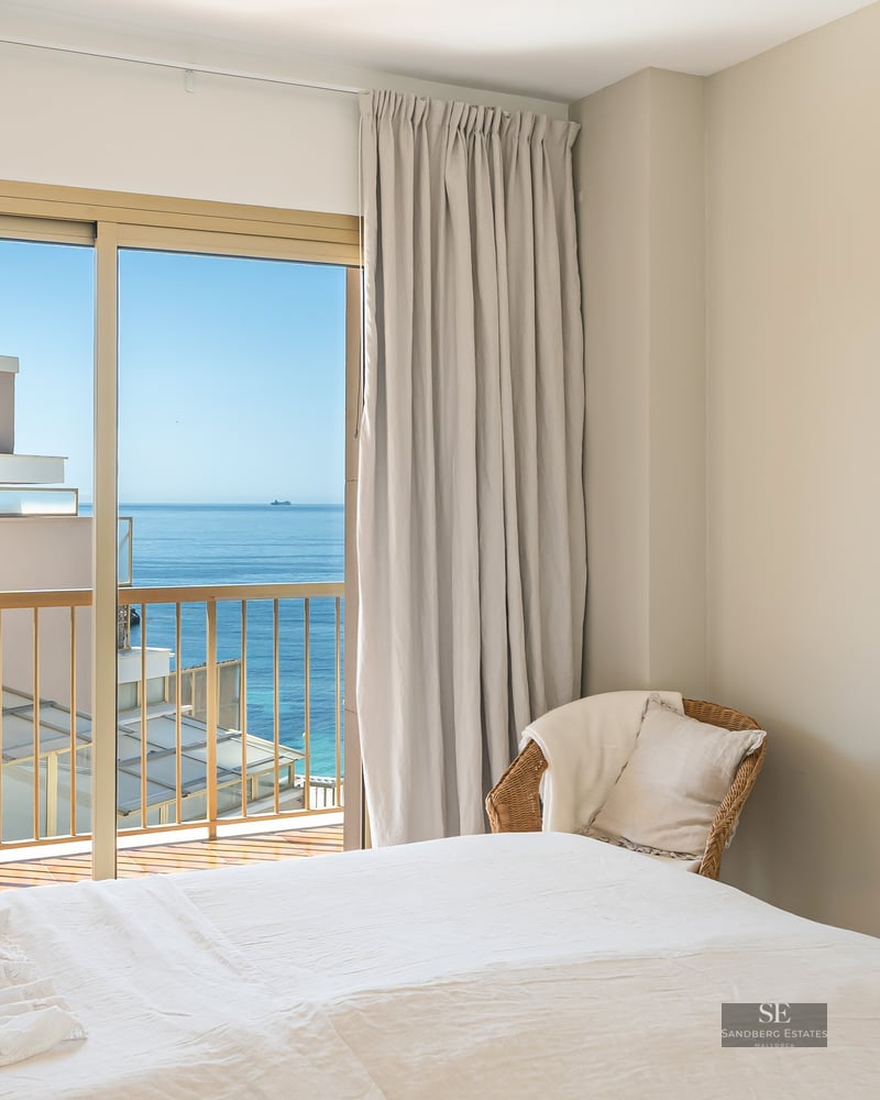 Sunlit bedroom featuring a white bed, wicker chair, and sliding glass doors opening to a sea view balcony.