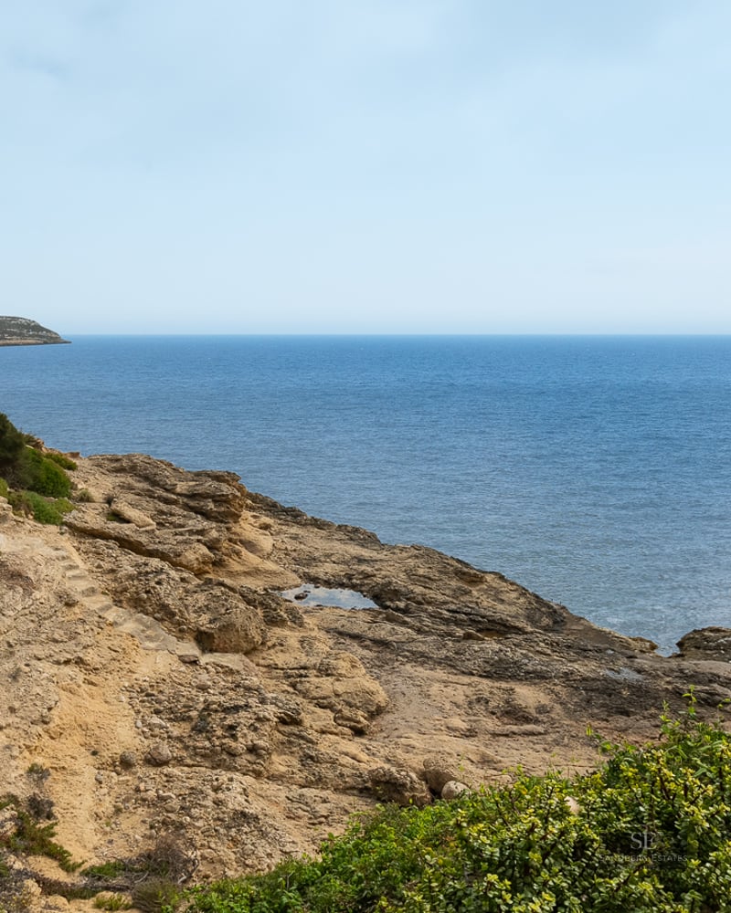Rocky coastline meeting the blue sea with green Mediterranean shrubs in the foreground.