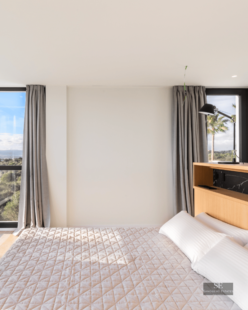Bright bedroom with large windows overlooking trees, wooden headboard with black marble, and white bedding.