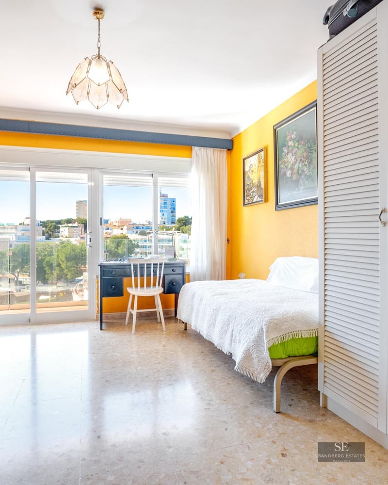 A sunny bedroom featuring yellow walls, a white slatted wardrobe, a desk by a large window, and city views.