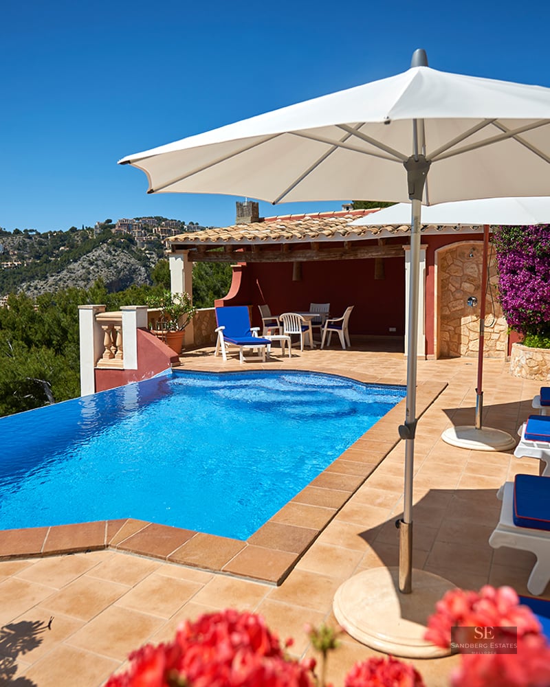 A bright blue swimming pool on a terracotta terrace with sun loungers and umbrellas overlooking hills and the sea.