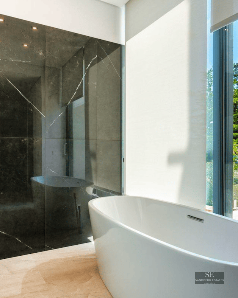 Modern master bathroom featuring a white freestanding bathtub next to a floor-to-ceiling window with green garden views.