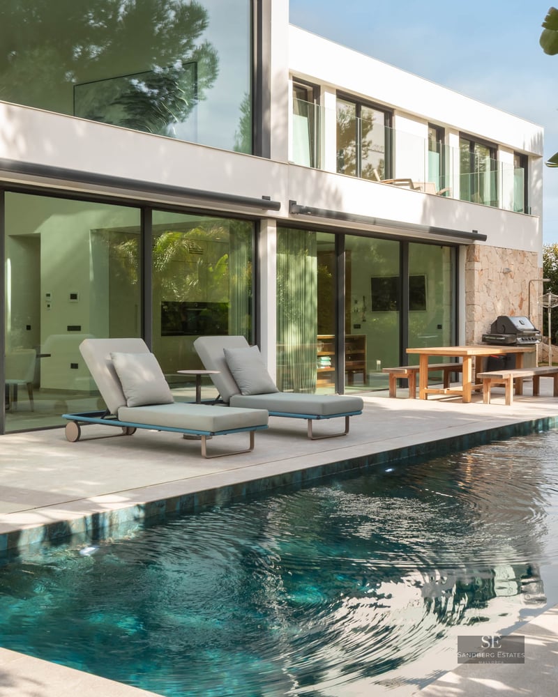 Piscine turquoise à côté d'une villa blanche moderne avec de grandes baies vitrées et des transats.