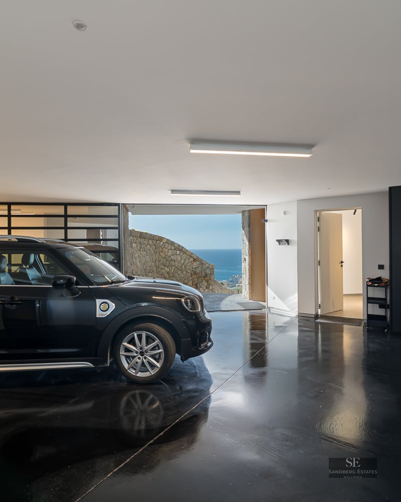 A black car parked in a modern garage with a glossy black floor, EV charger, and a sea view in the background.