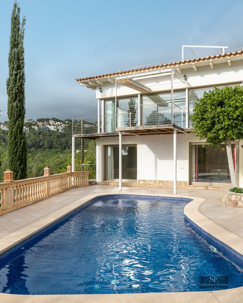 A kidney-shaped swimming pool on a stone terrace next to a two-story white villa overlooking a lush green forest.
