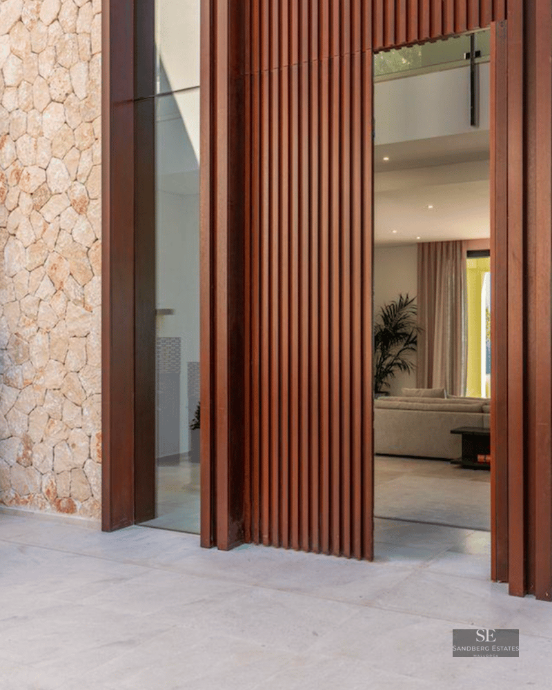 Grand villa entrance featuring a massive slatted wood door, natural stone walls, and spiral topiary in white planters.