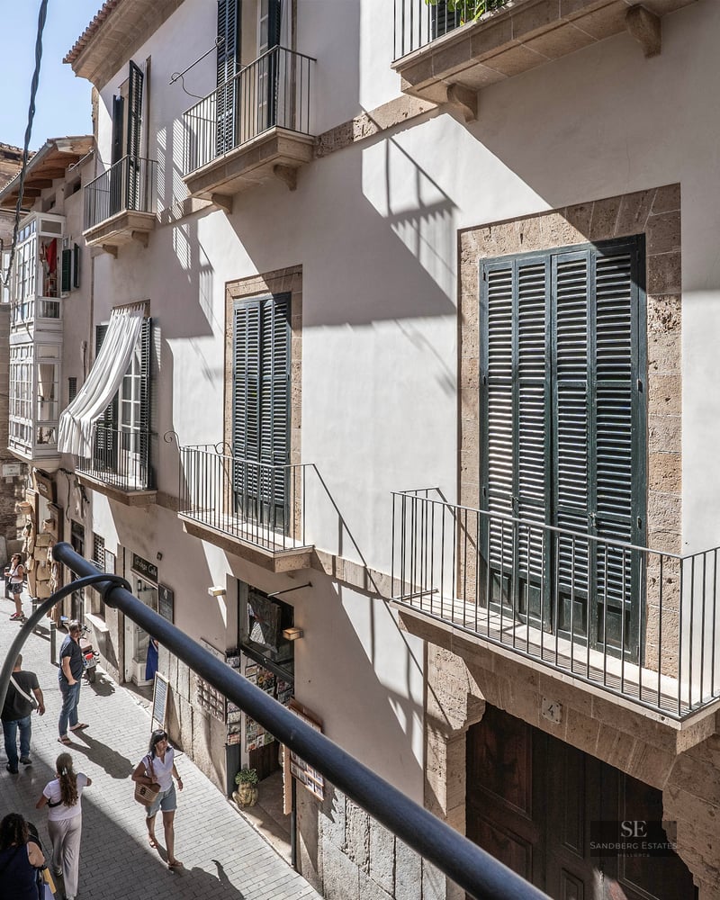 View from a balcony looking down a narrow street with light stone buildings, green shutters, and people walking.