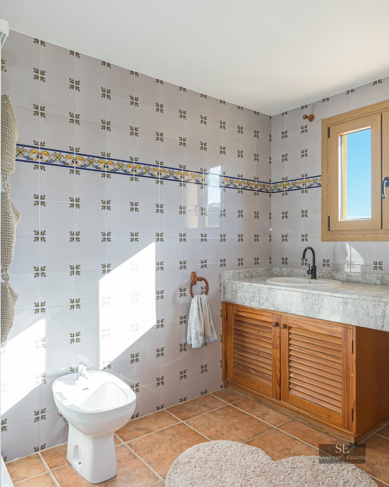 Bright bathroom featuring decorative wall tiles, white marble countertop, wooden vanity, bidet, and terracotta flooring.