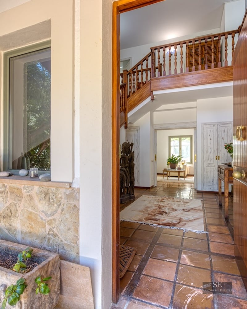 Open wooden door looking into a Mediterranean hallway with terracotta tiles and a wooden staircase.