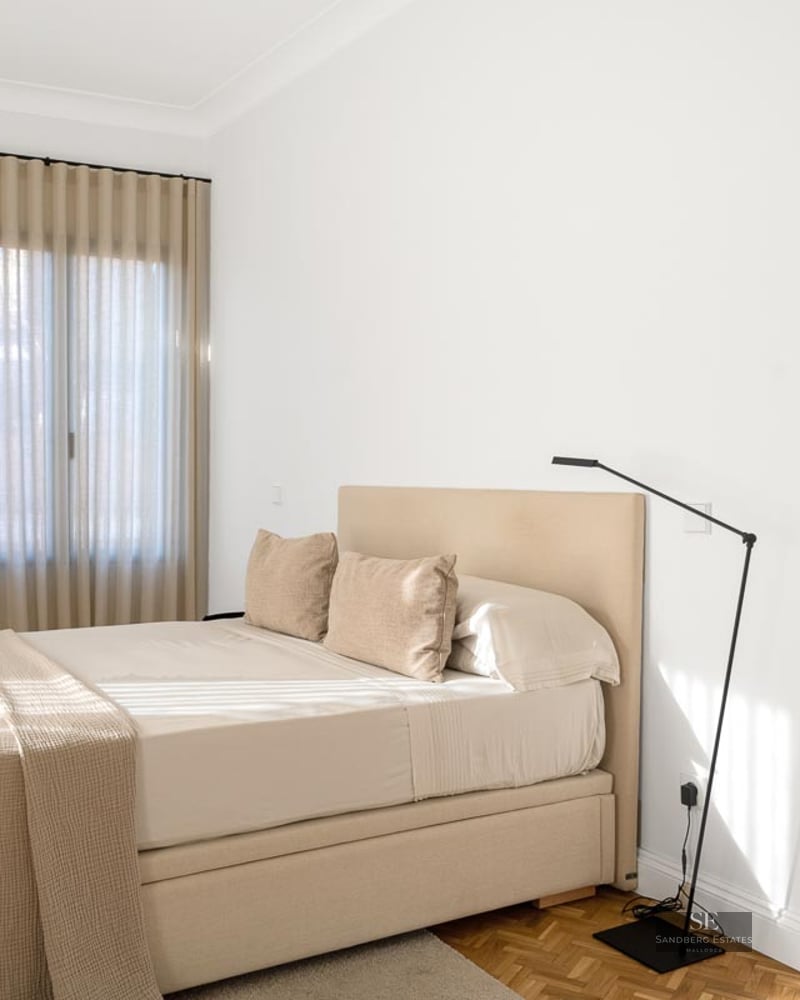Bright minimalist bedroom with a beige upholstered bed, white walls, and warm wooden parquet flooring.