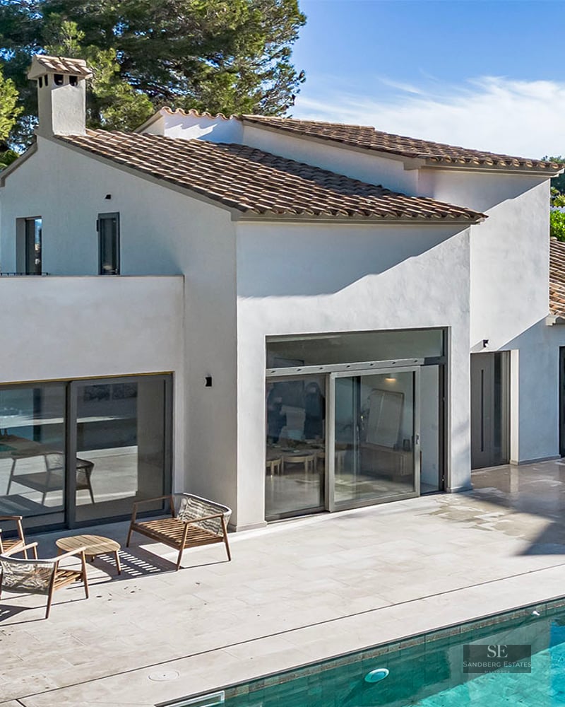 Modern white villa with a terracotta roof, large glass doors, and a stone terrace next to a turquoise swimming pool.