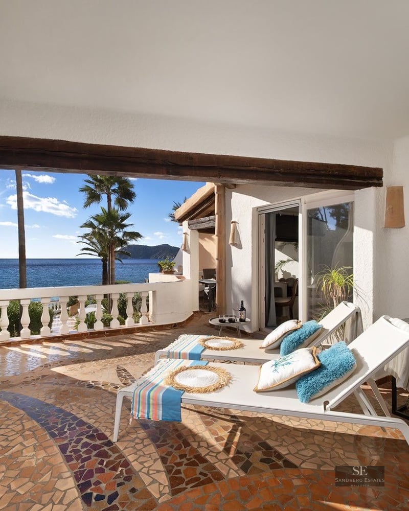 Sunlit terrace with mosaic floors, white loungers, and a stunning view of the blue ocean and palm trees.