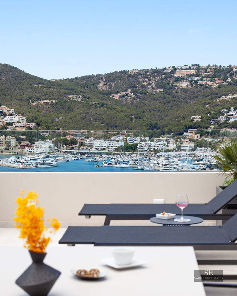 Sun loungers on a modern terrace overlooking a harbor with white boats and lush green hills under a clear blue sky.